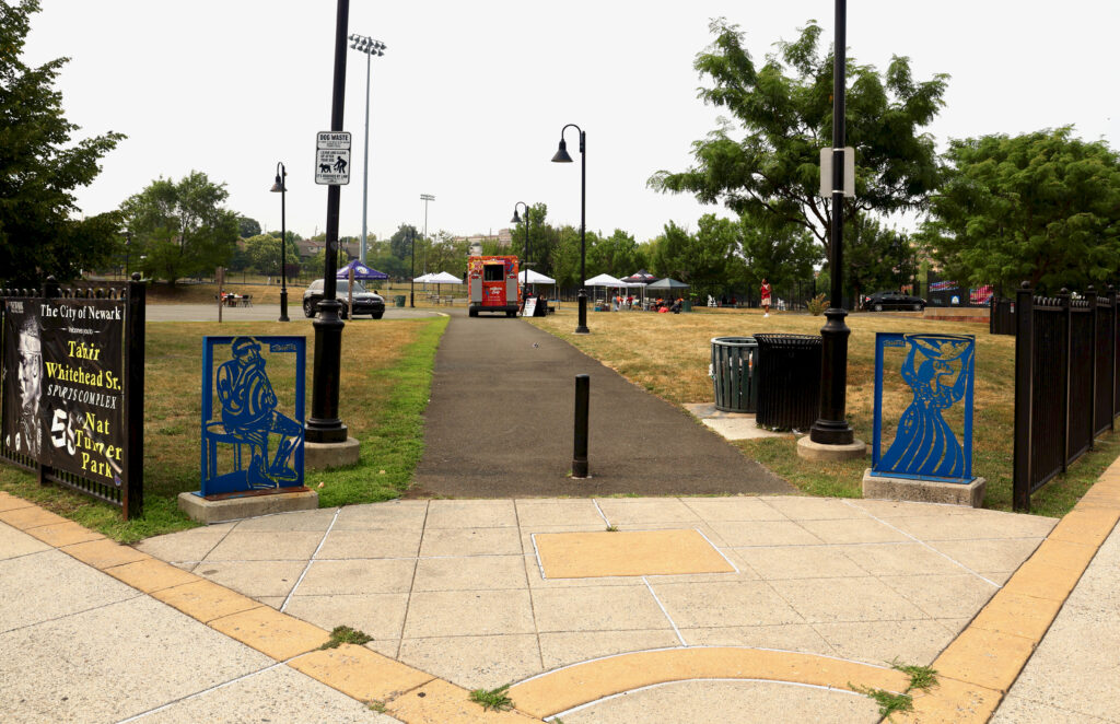 Entrance to Nat Turner Park in Newark featuring colorful steel art panels, walking path, and community event tents in the distance.